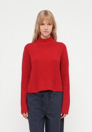 Red ribbed sweater with a high neck and long sleeves, featuring a relaxed fit and cropped hem. Paired with navy pinstriped trousers.