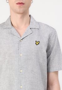 Kurzärmeliges, hellgraues Hemd mit Knopfleiste aus strukturiertem Stoff, mit einem gelben, gestickten Vogel-Logo auf der linken Brust.