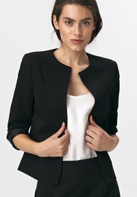 Kobieta w czarnym żakiecie na białej bluzce, trzymająca rękami brzegi żakietu w tle o jednolitej białej barwie.