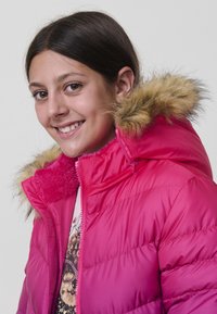 Chica sonriente con cabello oscuro que lleva una chaqueta de plumas rosa brillante con capucha forrada de piel y una camiseta estampada debajo, contra un fondo liso.
