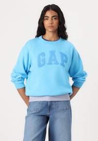 Sudadera azul claro con "GAP" en letras en relieve en la parte frontal. Presenta un cuello redondo y un corte oversized, combinada con jeans de mezclilla azules.