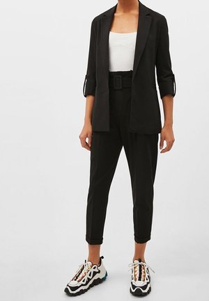 Femme portant un blazer noir aux manches retroussées, un pantalon noir ceinturé, un débardeur blanc et des baskets noires et blanches avec des détails colorés.