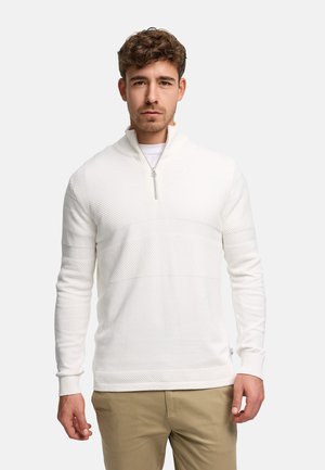 Man med kort brunt hår, klädd i vit zip-up-tröja med struktur och beige byxor, stående mot en enkel ljus bakgrund.