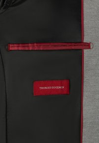 Tissu noir et gris avec des accents rouges ; présente une poche zippée et une étiquette avec "THOMAS GOODWIN" en blanc sur un fond rouge.