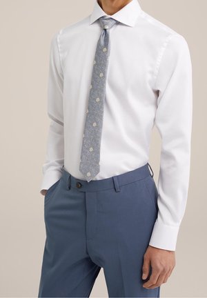 Mann trägt ein weißes Hemd, eine blaue Krawatte mit Blumenmuster und blaue maßgeschneiderte Hose, eine Hand in der Tasche, neutraler Hintergrund.