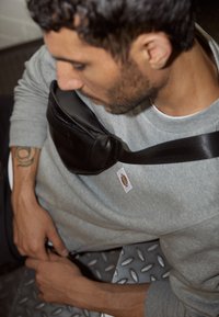 Borsa a tracolla in pelle nera con una tracolla testurizzata, indossata su una felpa grigia con un'etichetta tessuta. Sono visibili il braccio e una superficie metallica decorata.