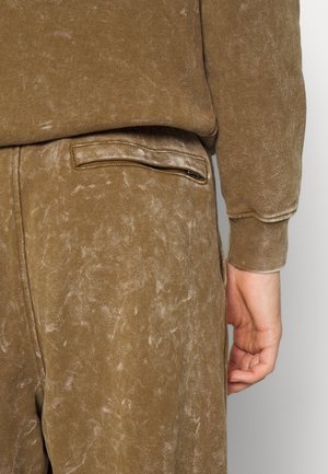 Detailní záběr na osobu nosící hnědé kalhoty s efektem acid wash a odpovídající vršek, zobrazující zadní kapsu a pravou ruku spočívající podél těla.