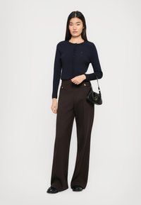 Cardigan cropped bleu marine avec fermeture à boutons, associé à un pantalon marron taille haute avec des accessoires en ton doré et des jambes larges, complété par un sac noir.