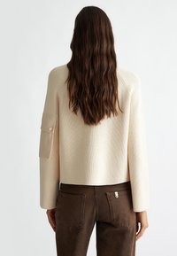 Pull en maille côtelée crème avec une coupe carrée et des manches raglan. Caractérisé par un détail de poche beige sur le bras gauche. Associé à un pantalon marron.