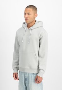 Gaiši pelēks kapucis izgatavots no mīksta auduma, ar priekšējo kabatu, kapuci ar aukliņu un ribotām aprocēm. Uz krūtīm ir smalki printēts logo.