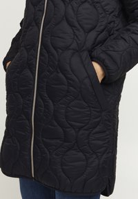 Chaqueta acolchada negra con patrón de olas, cremallera plateada, bolsillos laterales y puños acanalados. Textura suave, diseño hasta la rodilla.