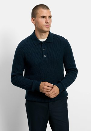 Homme aux cheveux courts portant un pull en maille bleu foncé avec col et boutons, debout sur un fond blanc, les mains jointes.