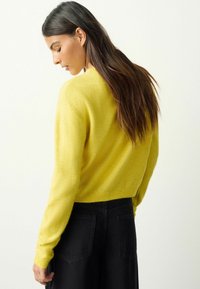 Kuvassa näkyy keltaisesta neuloksesta valmistettu pusero, jossa on pitkät hihat ja pyöreä pääntie; sen pehmeä materiaali näkyy. Pusero on yhdistetty korkeavyötäröisiin mustiin housuihin, taustakuvakulma.