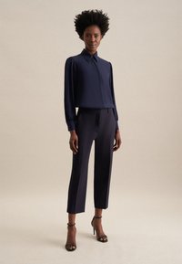 Blusa blu navy con colletto e maniche voluminose abbinata a pantaloni navy tagliati e su misura. La modella indossa sandali neri con tacco.