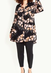 Blouse noire avec motif abstrait pierre orange et blanche, manches longues, col, associée à un pantalon noir slim.