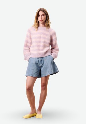 Vrouw staand met handen in de zakken, gekleed in een roze gestreepte, zachte trui, blauwe denim shorts en gele platte schoenen tegen een witte achtergrond.