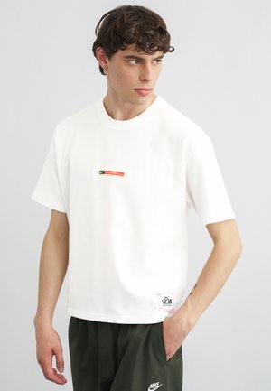 Mladý muž s krátkymi kučeravými vlasmi, oblečený v bielom Nike futbalovom tričku a tmavozelených Nike nohaviciach, pozerá sa doľava.