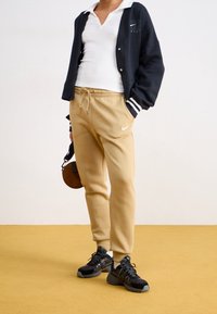 Czarny kardigan z białymi paskami, biała koszulka polo, beżowe spodnie dresowe i czarne buty sportowe. Mała brązowa okrągła torebka. Żółte tło.