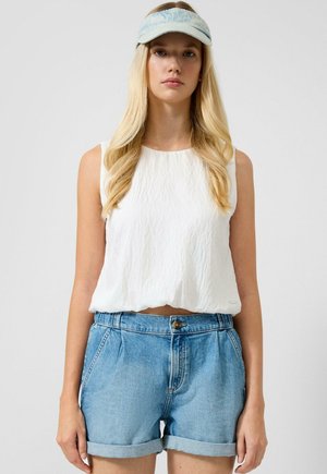 Junge Frau mit langen blonden Haaren, die ein weißes ärmelloses Oberteil, blaue Jeansshorts und eine hellblaue Schirmmütze vor einem einfarbigen Hintergrund trägt.