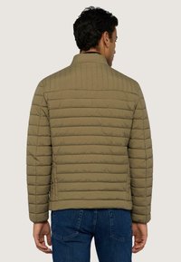 Bavlněná bunda ve vojenské zelené barvě, s horizontálním prošíváním, vysokým límcem a přiléhavým střihem, doplněná tmavými džíny.