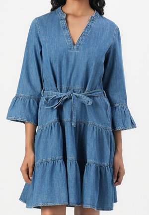 Kvinde iført en knælang blå denimkjole med 3/4 klokkeærmer, lagdelt nederdel, V-halsudskæring og bundet bæltet i taljen.