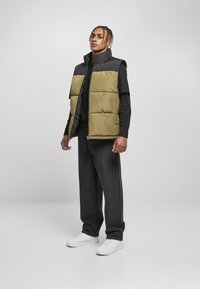 Polstret vest i oliven og sort, med høj krave, lynlås lukning og sidelommer. Båret over en sort langærmet skjorte og parret med mørke jeans.