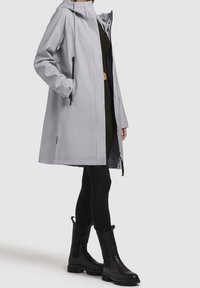 Personne portant un imperméable à capuche gris clair, des leggings noirs et des bottes noires mi-mollet, debout devant un fond gris uni.