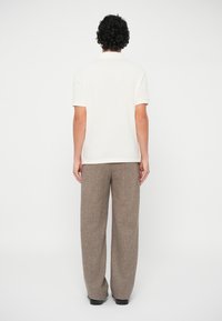 Άνδρας με σγουρά μαύρα μαλλιά, φοράει λευκό πόλο μπλουζάκι κοντομάνικο, καφέ φαρδιά παντελόνια και μαύρα παπούτσια, στέκεται γυρισμένος.