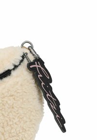 Bolsita de forro polar color crema con cremallera, que presenta una etiqueta negra con texto rosa. Material texturizado, con detalles en hardware plateado en la cremallera.