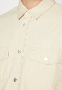 Short-sleeve beige shirt in lightweight fabric, featuring a button-down collar, silver buttons, and two front patch pockets.