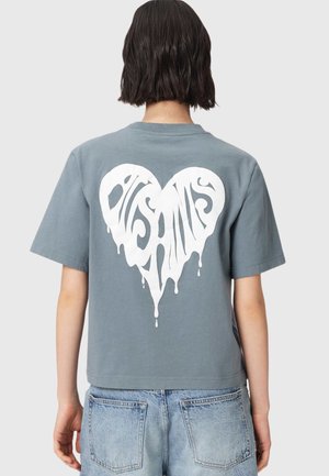 Person med kort mørkt hår iført en grå t-shirt med et stort hvidt dryppende hjertegrafik og blå jeans, set bagfra.