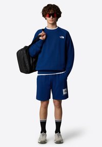 Sudadera azul y pantalones cortos, material de algodón, logo en el pecho y en la pierna, combinados con una bolsa negra, gafas de sol rojas y calzado gris con calcetas negras.