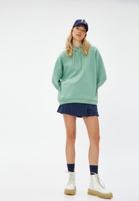 Sudadera verde menta de gran tamaño con cordones, combinada con una falda azul marino plisada y botas blancas con cordones y suelas de color canela.