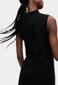 Schwarzes ärmelloses Kleid mit hohem Kragen und einem Reißverschluss am Rücken. Der Stoff wirkt glatt und hat eine figurbetonte Silhouette.
