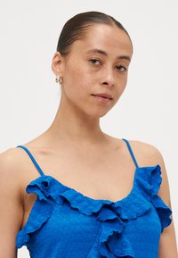 Plava ruffled majica s tankim naramenicama, koja ima teksturirani materijal. Model je upotpunio izgled malim kružnim naušnicama.