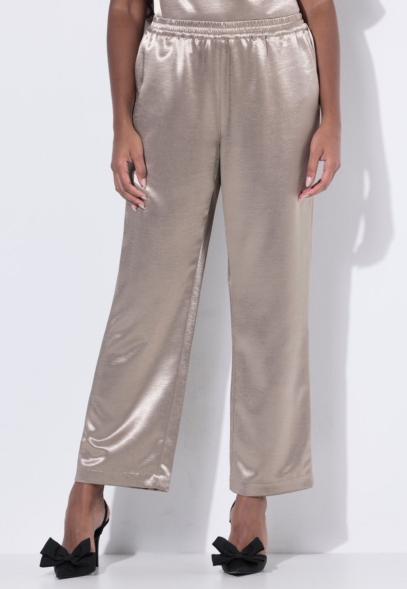 Donna con pantaloni beige larghi e lucidi e tacchi neri con grandi fiocchi, in piedi davanti a uno sfondo bianco uniforme.