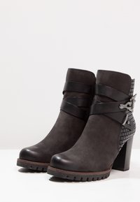 Par de botas de tobillo de ante gris oscuro con correas de envoltura negra, hebillas plateadas, tacón texturizado y suela gruesa, fotografiadas sobre fondo blanco.