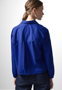 Camicia blu a maniche lunghe realizzata in tessuto morbido. Caratterizzata da un colletto, cuciture visibili e una vestibilità comoda con dettagli arricciati nella parte posteriore.