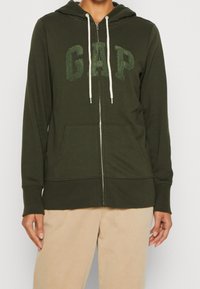 Sudadera con cremallera en color verde oliva, con un bolso delantero, que presenta cordones blancos y el logotipo texturizado de GAP en el centro. Material de algodón suave.