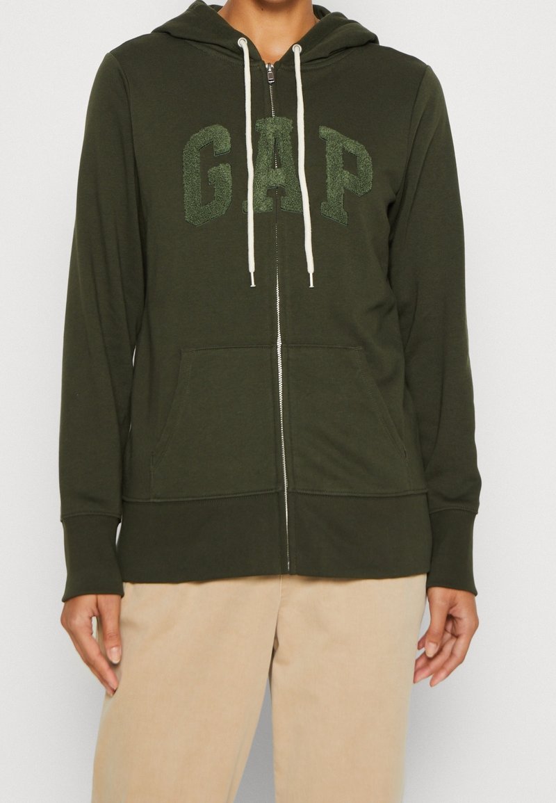 Sudadera con cremallera en color verde oliva, con un bolso delantero, que presenta cordones blancos y el logotipo texturizado de GAP en el centro. Material de algodón suave.