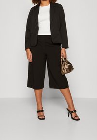 Blazer negro ajustado sobre una blusa blanca, combinado con pantalones culotte negros. Accesorios con un bolso metálico bronce y sandalias de tacón negras.