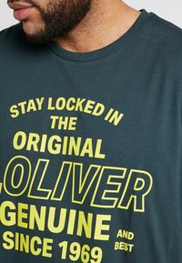 Donkerblauwe T-shirt met een ronde halslijn en korte mouwen, met grote gele tekst die zegt: "BLIJF GESLOTEN IN DE ORIGINELE OLIVER ECHT SINDS 1969."