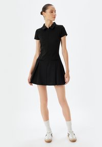 Vestido corto negro de manga corta con cuello, cierre de cremallera en la parte delantera y dos bolsillos frontales. Diseño de falda acampanada, textura suave y mínimos detalles.