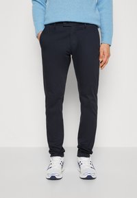 Pantaloni slim-fit navy in cotone misto con due tasche frontali e chiusura a bottone. Indossati con sneakers sportive bianche e un maglione azzurro chiaro.