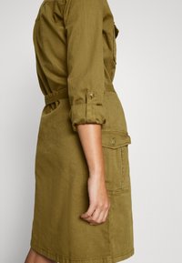 Abito utility verde oliva con maniche arrotolate, vita con cintura e dettagli di tasche frontali, realizzato in tessuto resistente con una texture liscia.