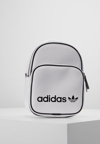 Bílý mini batoh Adidas s černým logem na přední kapse, postavený na bílém podstavci na pozadí bez vzorů. 