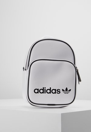 Biały mini plecak Adidas z czarnym logo na przedniej kieszeni, stojący na białym cokole na tle jednolitego jasnego tła.