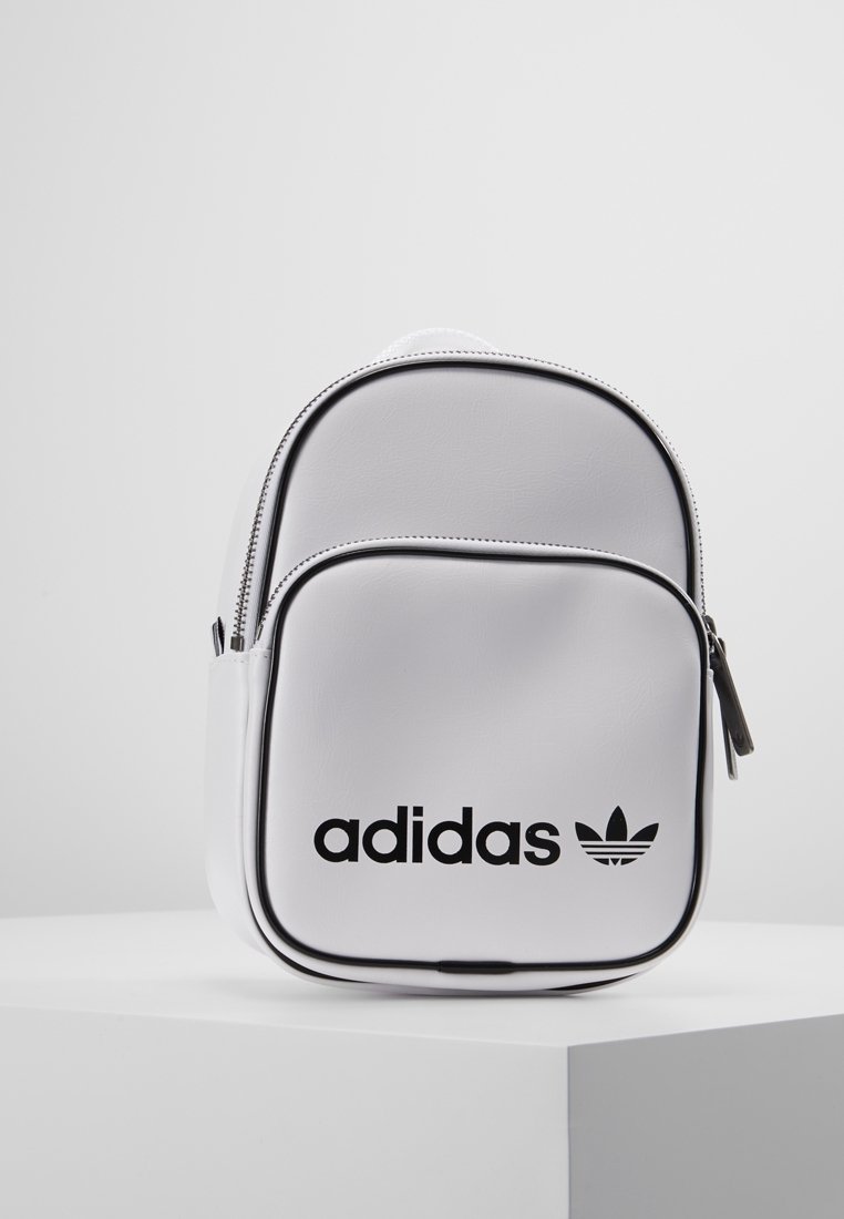 Bílý mini batoh Adidas s černým logem na přední kapse, postavený na bílém podstavci na pozadí bez vzorů. 