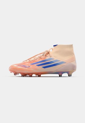 Chuteira de futebol com um design elegante, parte superior na cor pêssego, riscas azuis, superfície texturizada e cano alto. Sola com cravos para tração.