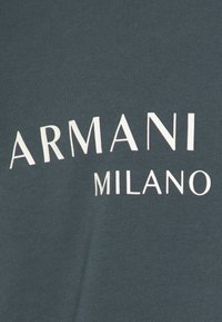 Camicia verde scuro con testo bianco che dice "ARMANI MILANO" in un design curvo. Il tessuto appare morbido e a coste.